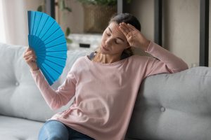Woman sitting on a couch fanning herself with a blue fan, indicating need for AC maintenance.