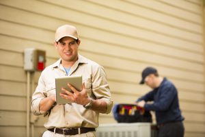 Technician using a tablet while another repairs an outdoor AC unit, representing AC repair and air conditioning repair services.