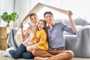 Smiling family sitting on the floor holding a cardboard roof, symbolizing home protection and emergency HVAC service.