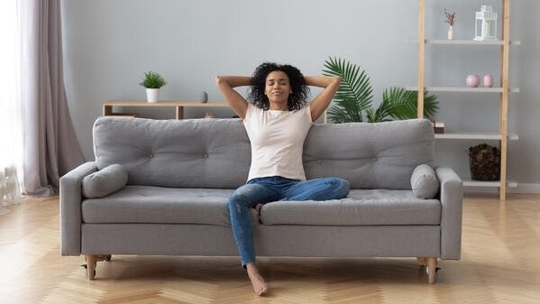 Relaxed woman sitting on a gray couch in a cool, air-conditioned living room, suggesting AC repair.